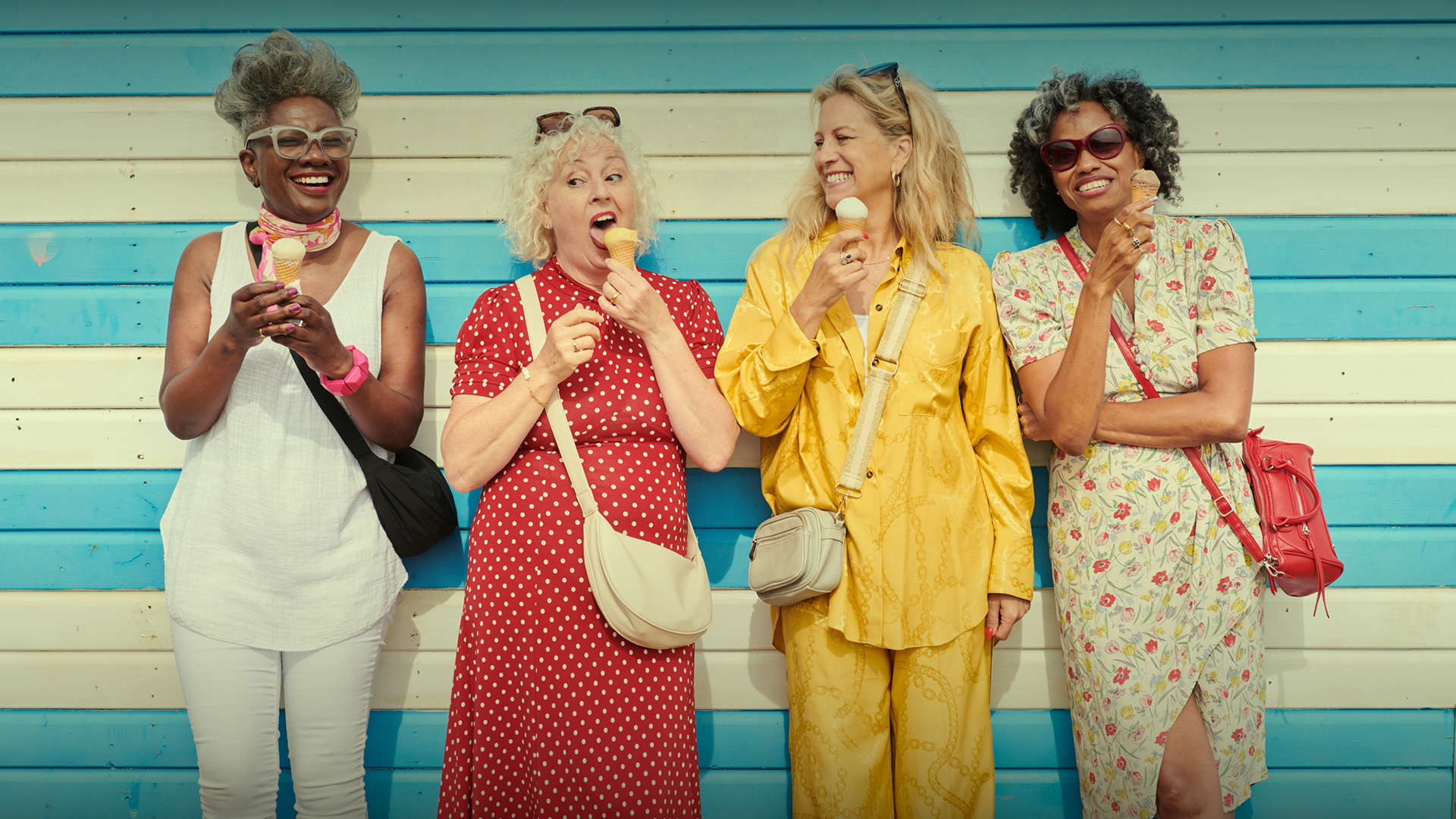 “Four older women smiling and enjoying ice cream while standing together in front of a blue striped wall on a sunny day.
